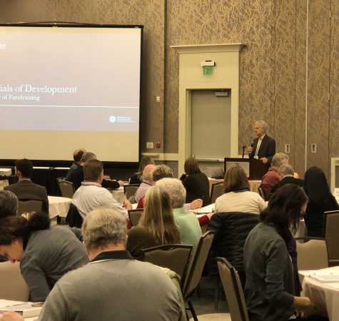 A man stands at a podium giving a presentation in front of an audience sitting around tables.