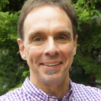 A man with short brown hair with gray facial hair wearing a checkered shirt smiles at the camera.