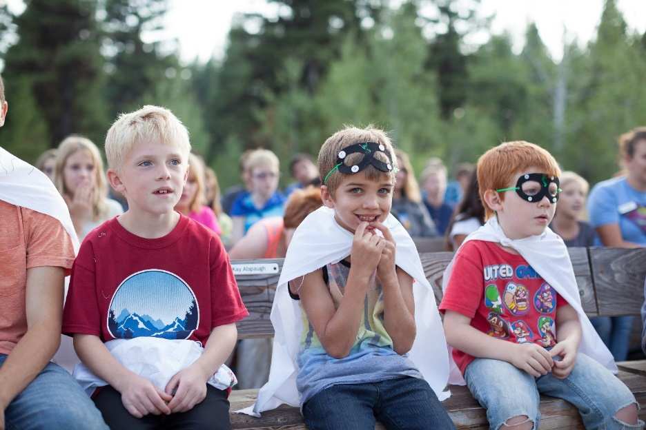 Three young boys, two of them wearing eye masks and white capes, sit on a wooden bench outside.