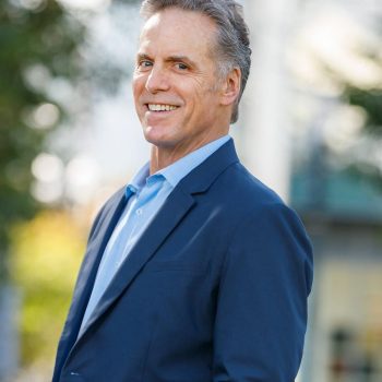 A man with gray hair wearing a blue shirt and suit smiles for the camera outside.