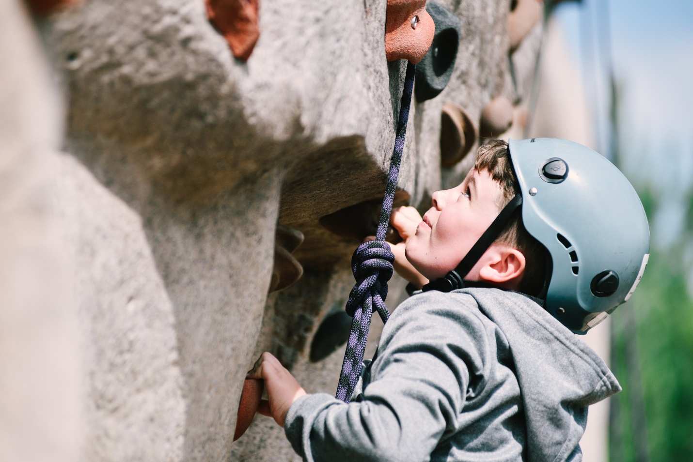 A young boy wearing a blue helmet and gray sweatshirt climbs a rock wall.