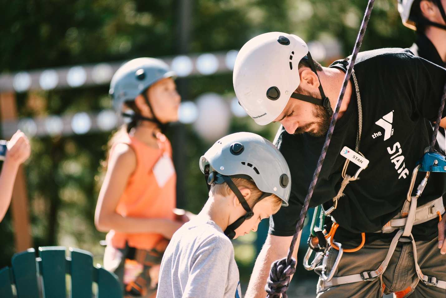 A man wearing a white helmet and black t-shirt helps a young boy wearing a blue helmet and gray t-shirt outside.