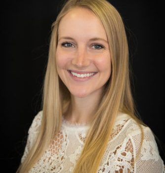 A young woman with long, straight blond hair wearing a white shirt smiles at the camera in front of a black background.