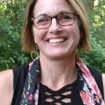 A woman with short brown hair wearing glasses, hoop earrings, a black shirt, and a scarf smiles at the camera.