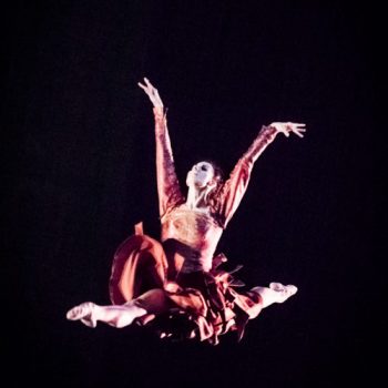 A ballerina wearing a pink dress performs a leap mid-air against a black background.