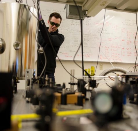 A man with short dark hair wearing dark glasses and a black jacket works in a laboratory.