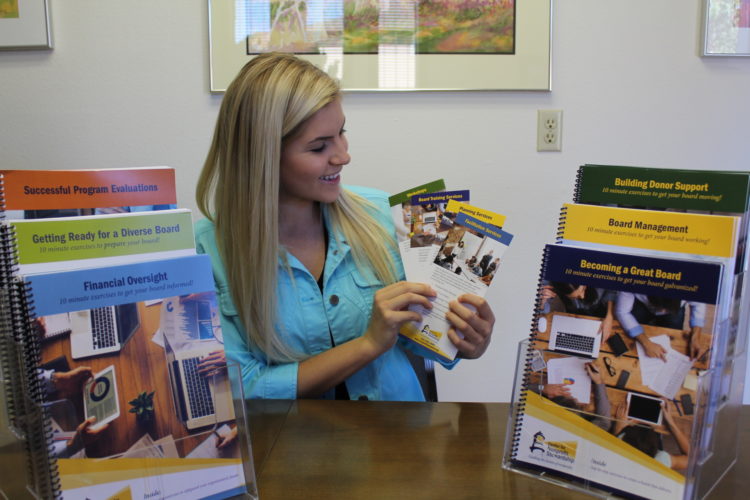 A woman with long, straight, blond hair smiles while holding pamphlets and sitting at a table.