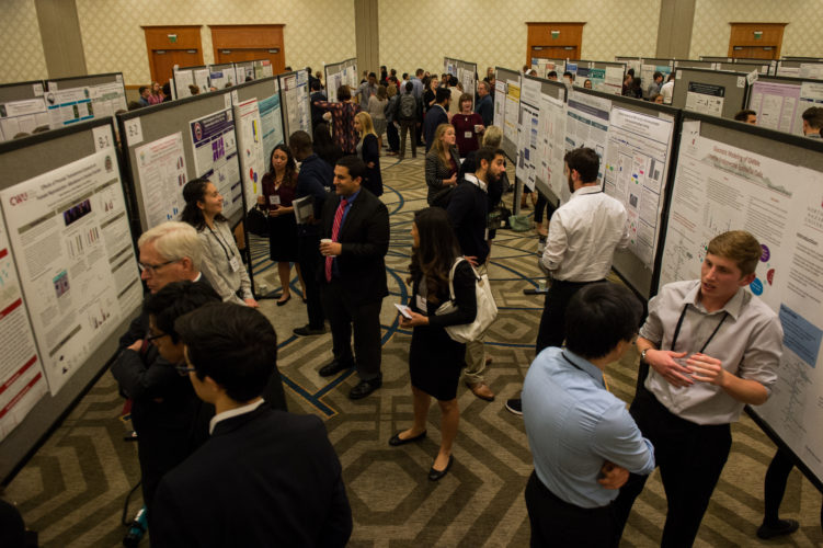 A group of people inside a large room having conversations in front of large posters with presentations on them.