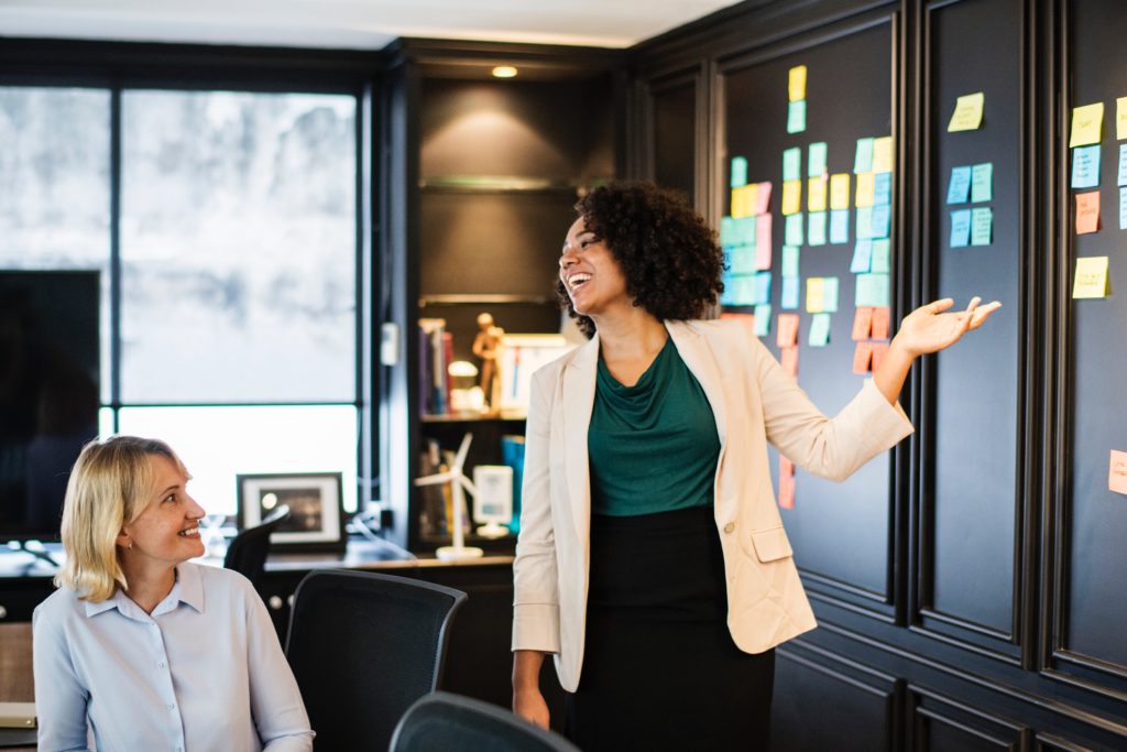 Two women have a lively conversation inside an office space with sticky notes on the wall.