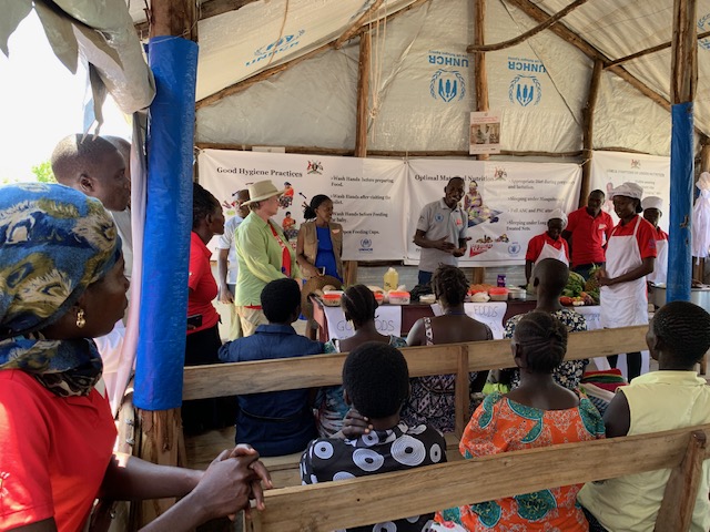 Over a dozen people gather inside a large tent to receive instruction on health, wellness, and nutrition at a Ugandan refugee camp.