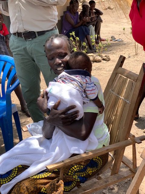 A woman with dark hair and skin sits outdoors in a rocking chair while holding a baby to her chest.