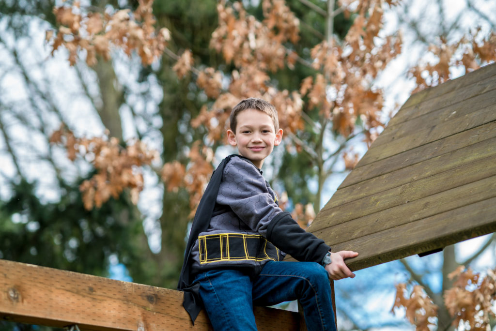 A young boy sits on a wooden beam with trees in the background and smiles at the camera.