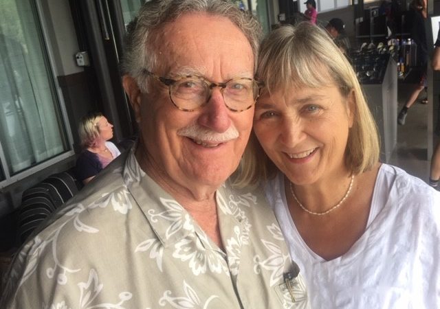 A man with gray hair and a mustache wearing a tan and white Hawaiian shirt and glasses smiles at the camera next to a woman with short blond hair and bangs wearing a white shirt and necklace.