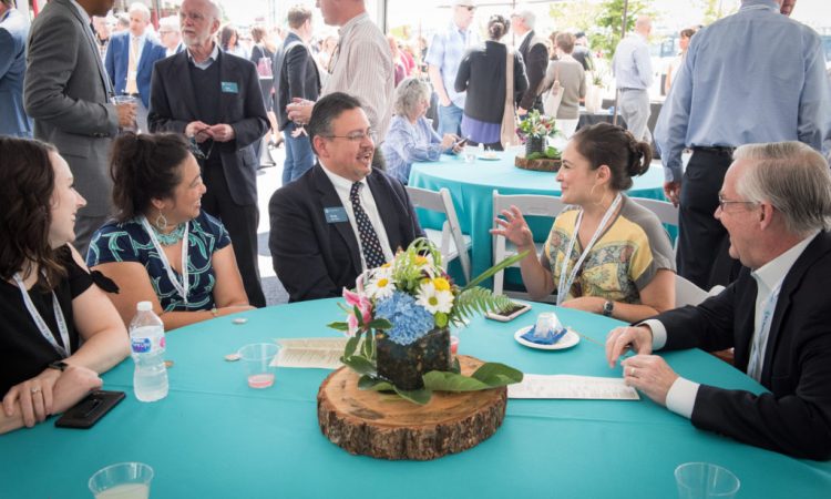 Five adults have a conversation while sitting around a circular table with blue tablecloth. A flower centerpiece is in the middle of the table. There are other blue tables and many other adults talking in the background.