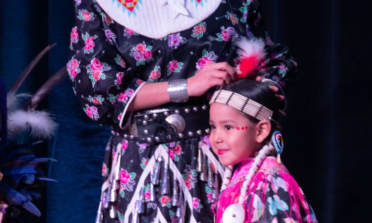 A young girl wearing a headdress, pink lipstick, and a bright pink fringed dress smiles at something off camera. A woman wearing a black dress with a pink rose pattern stands behind her.