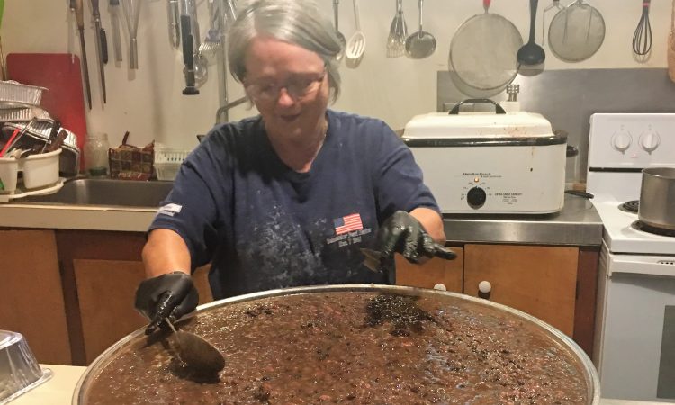 A woman with short gray hair and glasses wearing a blue t-shirt stirs a large pot of food inside a kitchen while wearing black gloves.