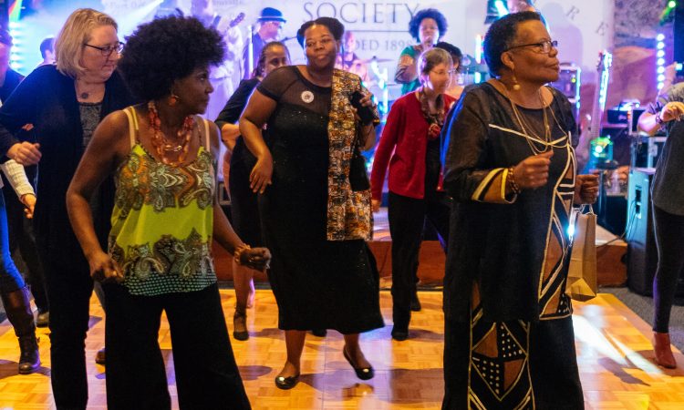 A group of women dance on a dance floor with a band playing in the background.