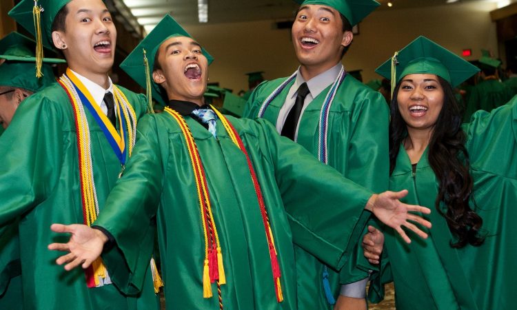 Three male and one female student wearing green graduation gowns and caps look excited for the camera.