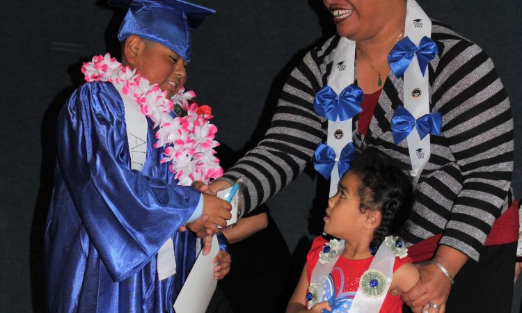 A young student wearing a blue graduation gown and cap and pink and white floral lei shakes hands with an adult. A small child stands by the adult and looks up.