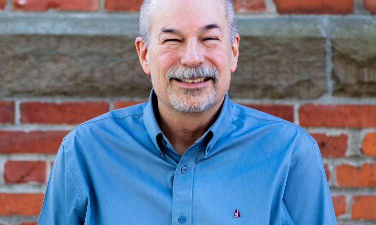 A man with gray hair and facial hair wearing a blue shirt smiles at the camera in front of a red brick wall.