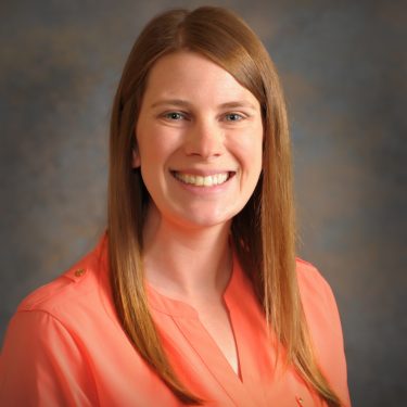 A woman with straight red hair wearing an orange shirt smiles for the camera.