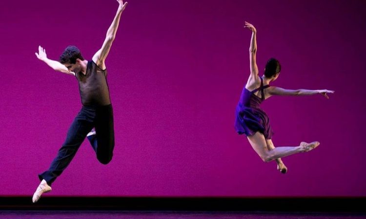 A male and female dancer perform a leap on stage.