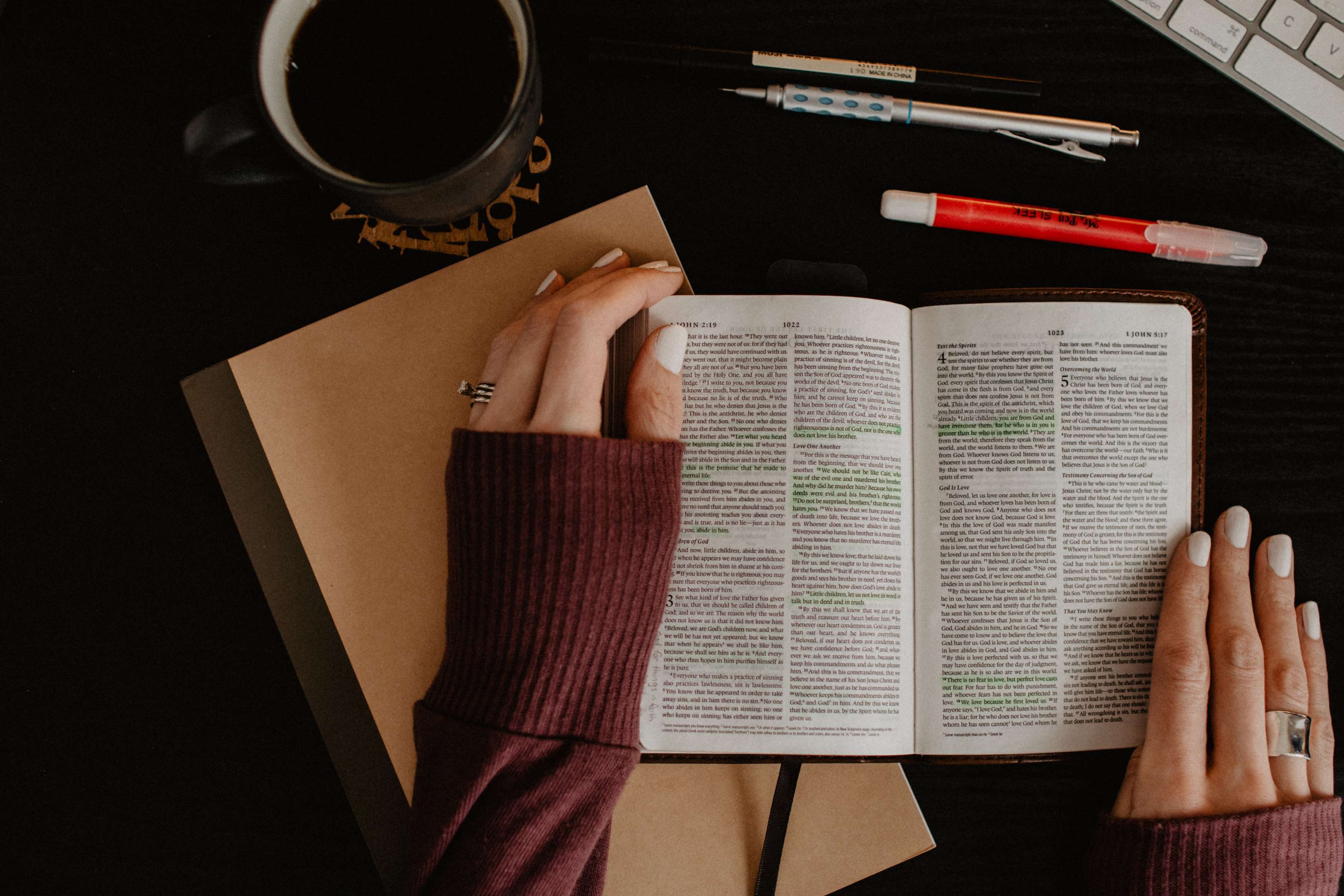 Two hands holding open a Bible on a table, with a notebook, pens, and coffee mug nearby.