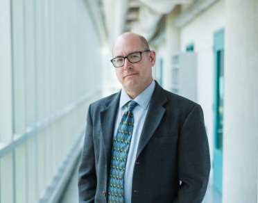 A man in a suit and glasses standing in a hallway looking at the camera.