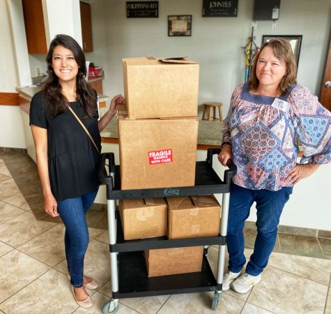 A woman with straight dark hair wearing a black shirt and jeans and a woman with straight brown hair wearing a patterned shirt and jeans smile for the camera next to a cart stacked with cardboard boxes.