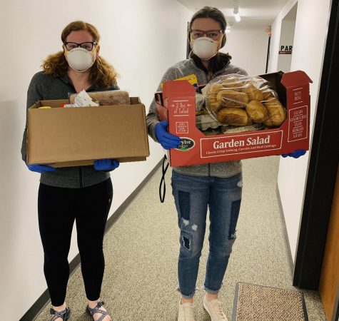 Two people wearing face masks and blue gloves stand in a hallway holding boxes of food.