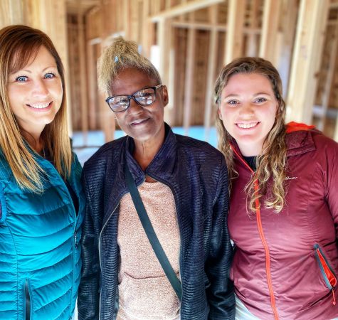 A woman with brown hair wearing a bright blue jacket, a woman with brown hair wearing a pink sweater and dark jacket, and a woman with brown hair wearing a pink jacket smile for the camera inside a construction site.