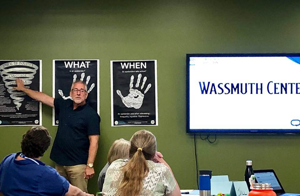 A man wearing glasses and a black shirt points to a poster on a wall inside a classroom.