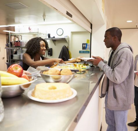 A woman with dark curly hair in an industrial kitchen hands a plate of food to a man with dark hair wearing a gray jacket.