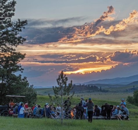 A group of people gather around a campfire, with a sunset in the background.