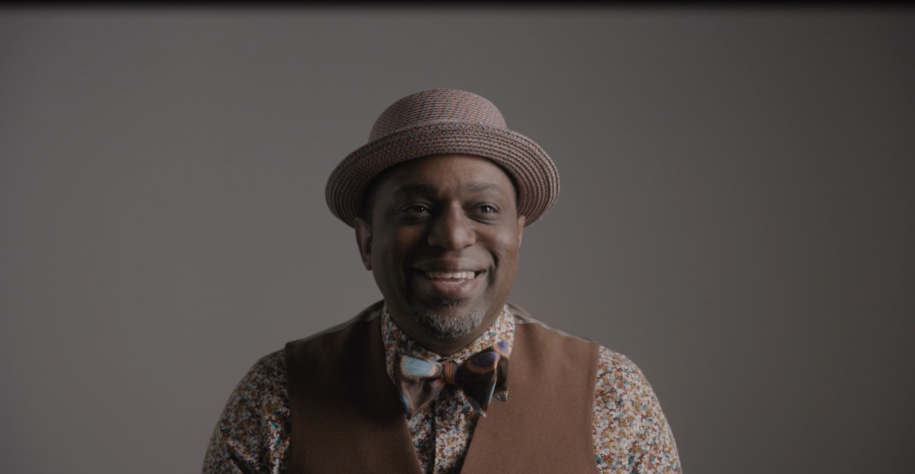 A man wearing a bowler hat, a vest, and a bowtie smiles in front of a dark background.