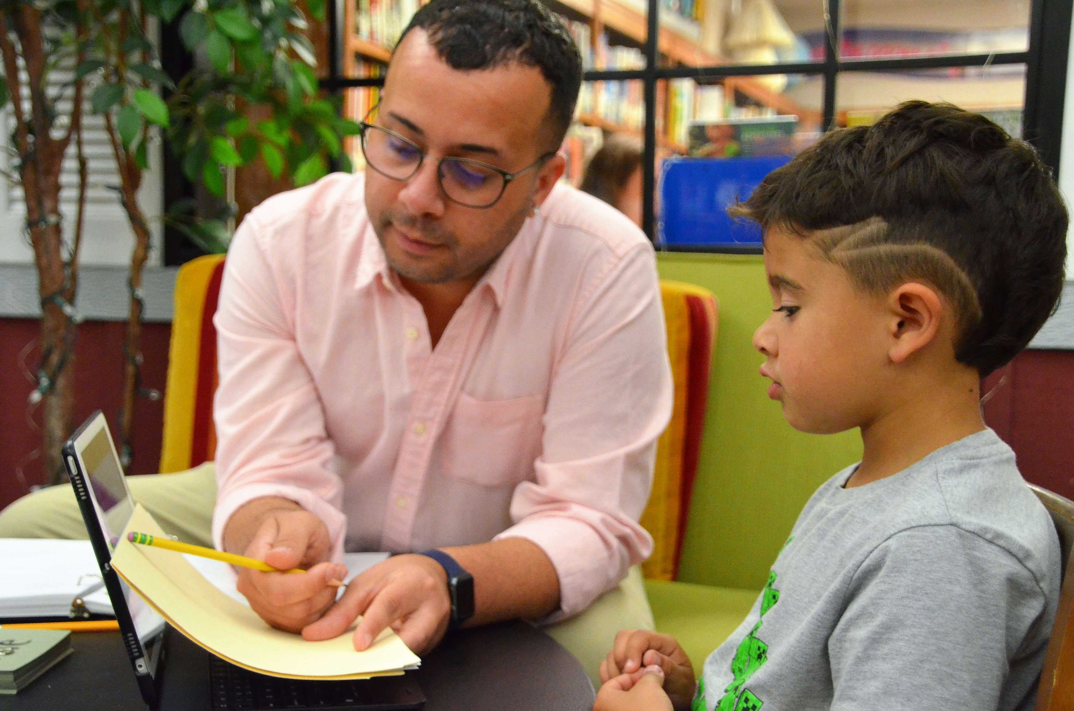 A man in a pink button-down shirt helps a young student learn to read.