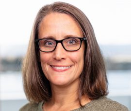 A woman with shoulder-length brown hair wearing glasses smiles at the camera.