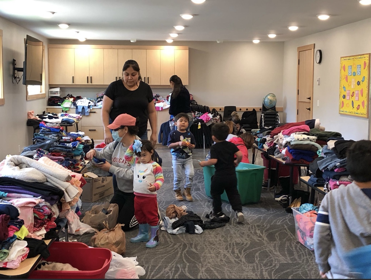 A room full of clothes and supplies with several adults and children sorting through them.