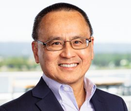 A man wearing glasses and a suit with a blue shirt smiles for the camera.