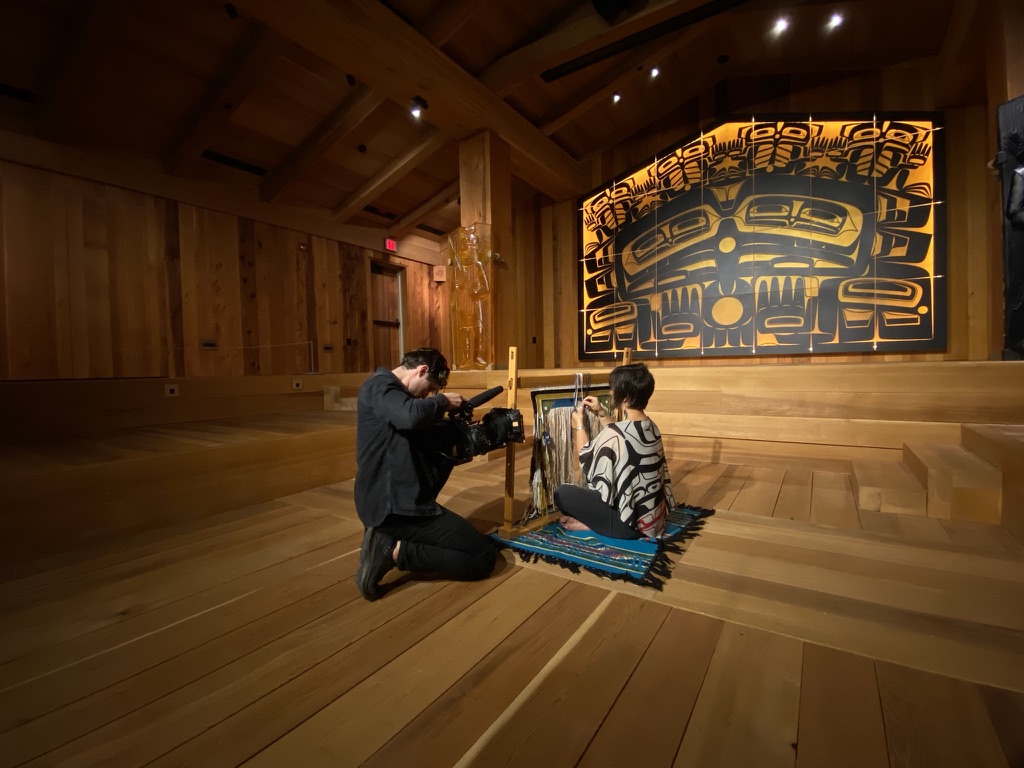 A cameraman takes a close up shot of a woman sitting on a blue woven mat, with a piece of Native American artwork in the background.