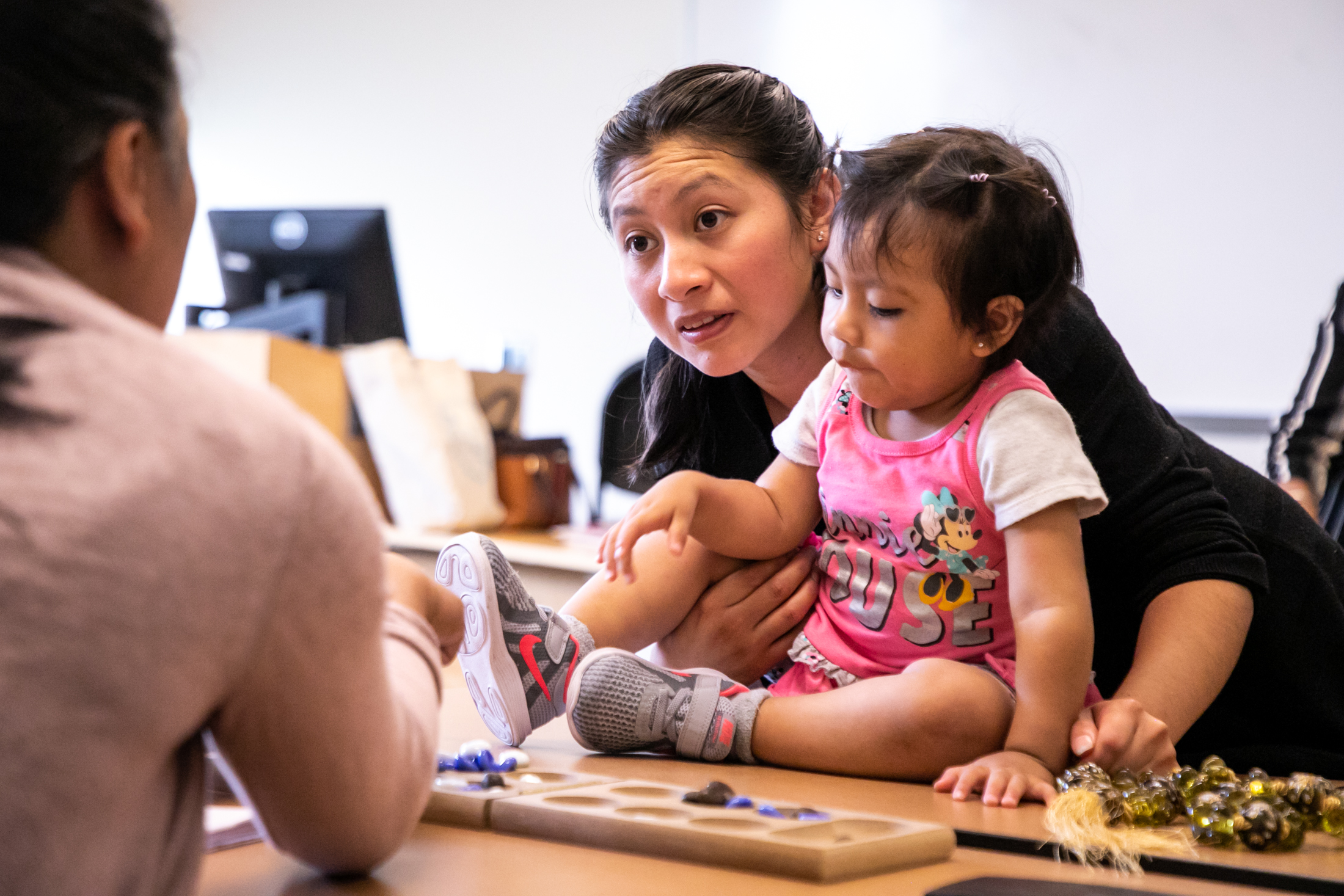 An educator with dark hair wearing black supports a young child with dark brown hair wearing a pink dress as she plays with marbles.