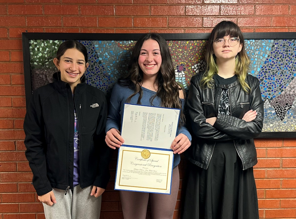 Three girls standing against a brick wall, with the one in the middle holding up an award