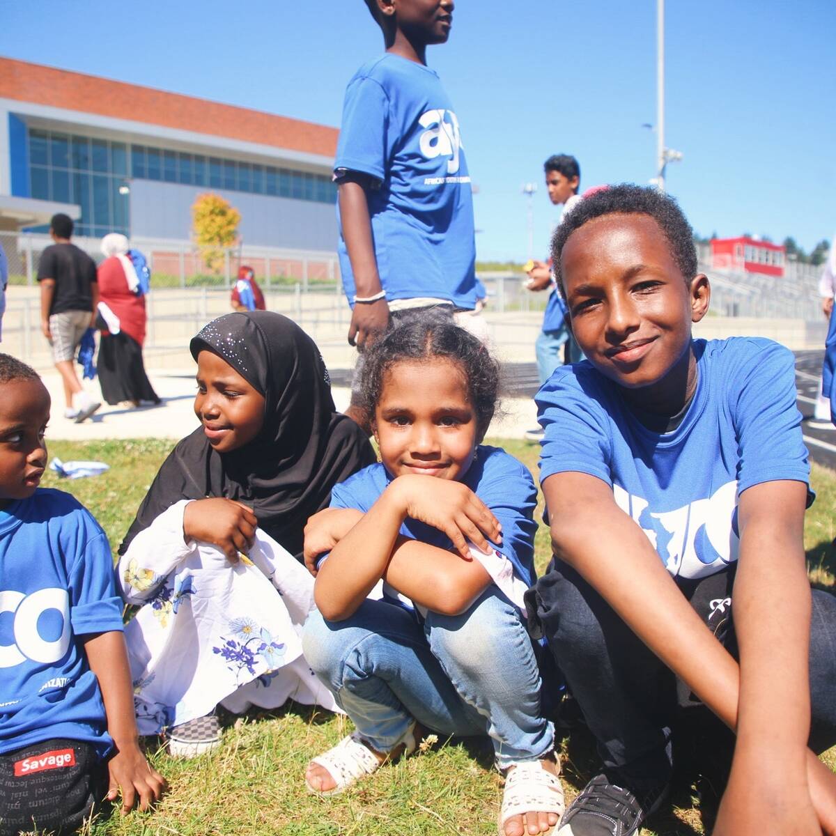Four kids sitting on grass outside a building with blue shirts that say AYCO (African Youth and Community Center)