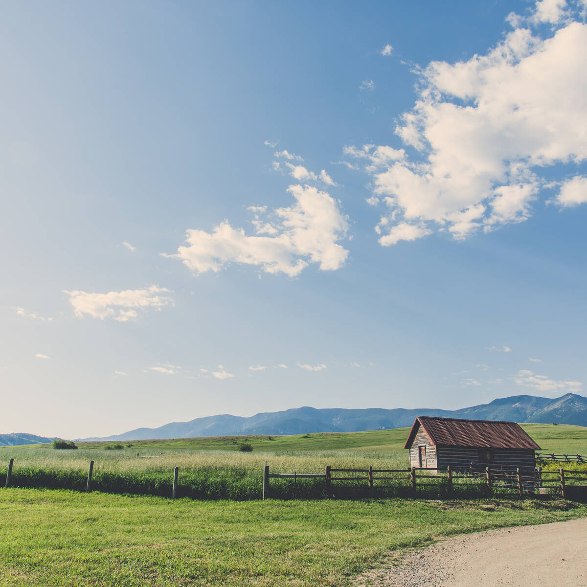 Scenic landscape photo of a green field in Montana with a small shed and wooden fence under a blue sky