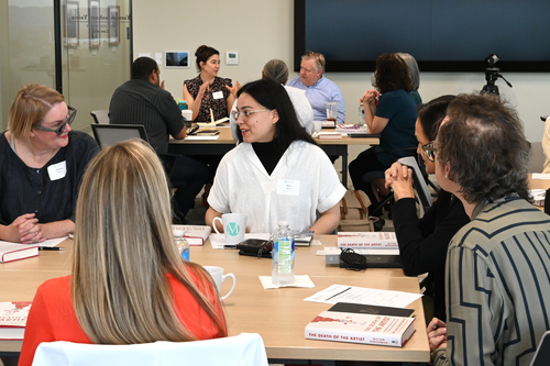 A group of five people have a conversation around a table, with another group at a different table behind them. They are inside a conference room with a TV in the background.
